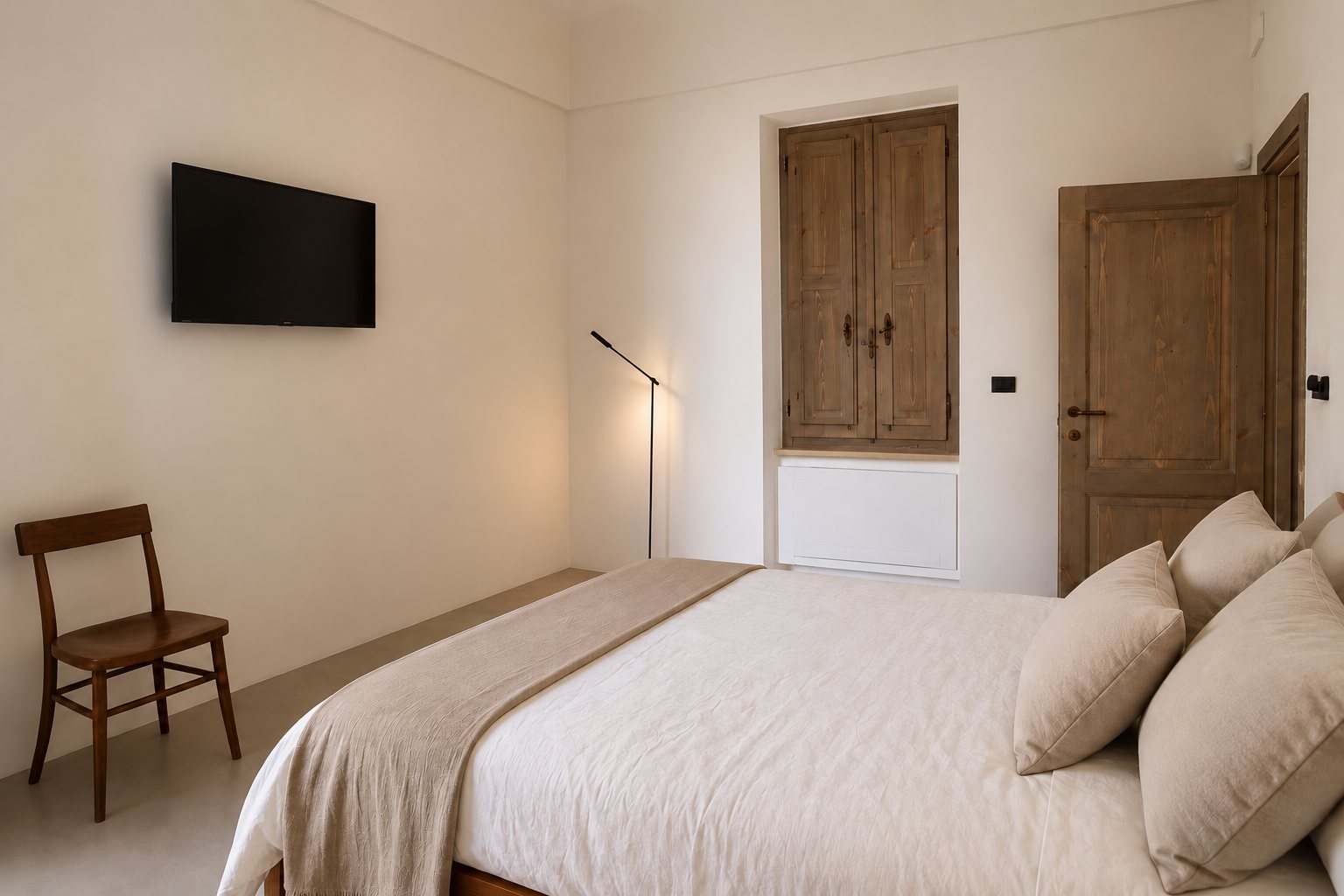 La seconde chambre du casale d'origine à Casa Andrea — des volets en bois ancien repliés dans l'embrasure, une porte ancienne en bois ouverte, un lit et ses coussins de lin, une chaise bistrot, un téléviseur mural, Pouilles.