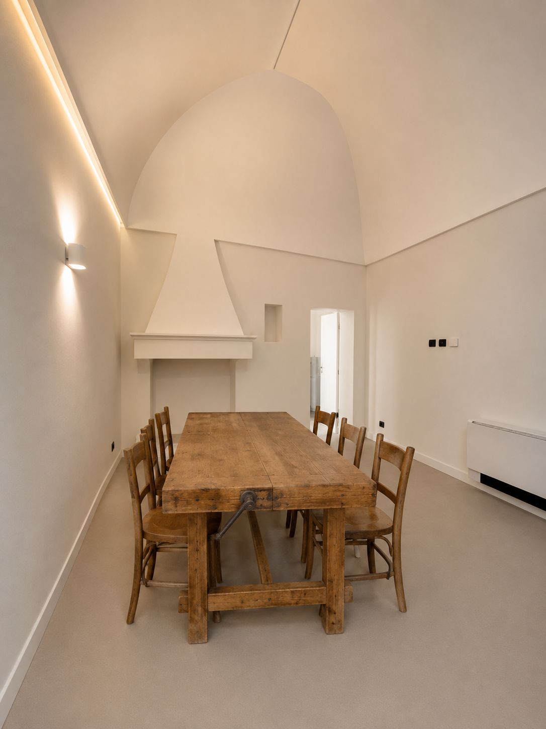 La salle à manger de Casa Andrea habitée — un ancien établi de menuisier chiné en brocante, devenu table de salle à manger, et ses chaises sous la voûte en coupole blanche, la cheminée conique en plâtre à côté, sol en microciment, Pouilles.