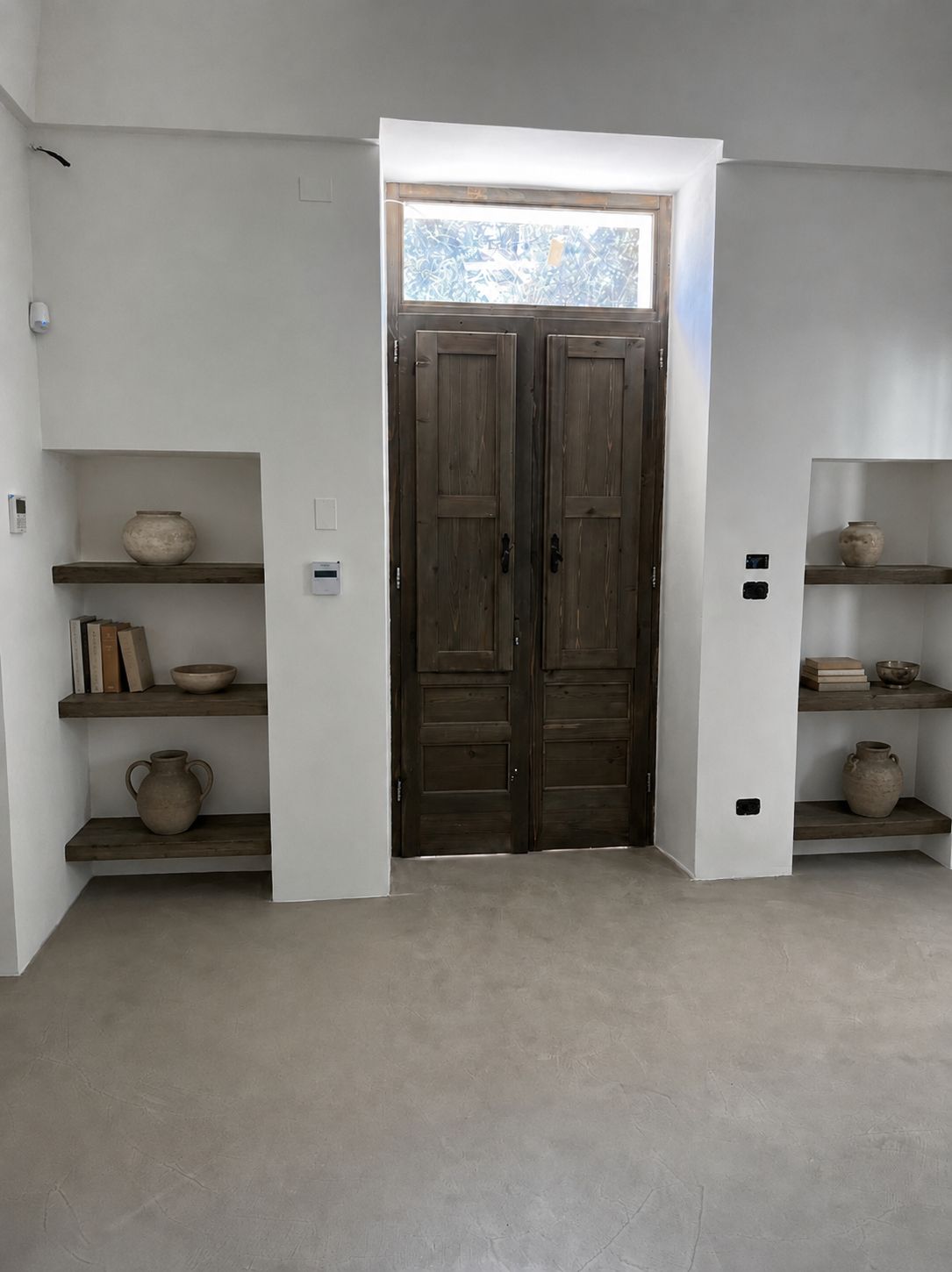 La porte d'entrée vue depuis la salle à manger — un ancien double battant en bois restauré avec une imposte en majolique peinte, flanqué de deux niches à étagères de bois garnies de jarres en terre cuite, sol en microciment, Pouilles.