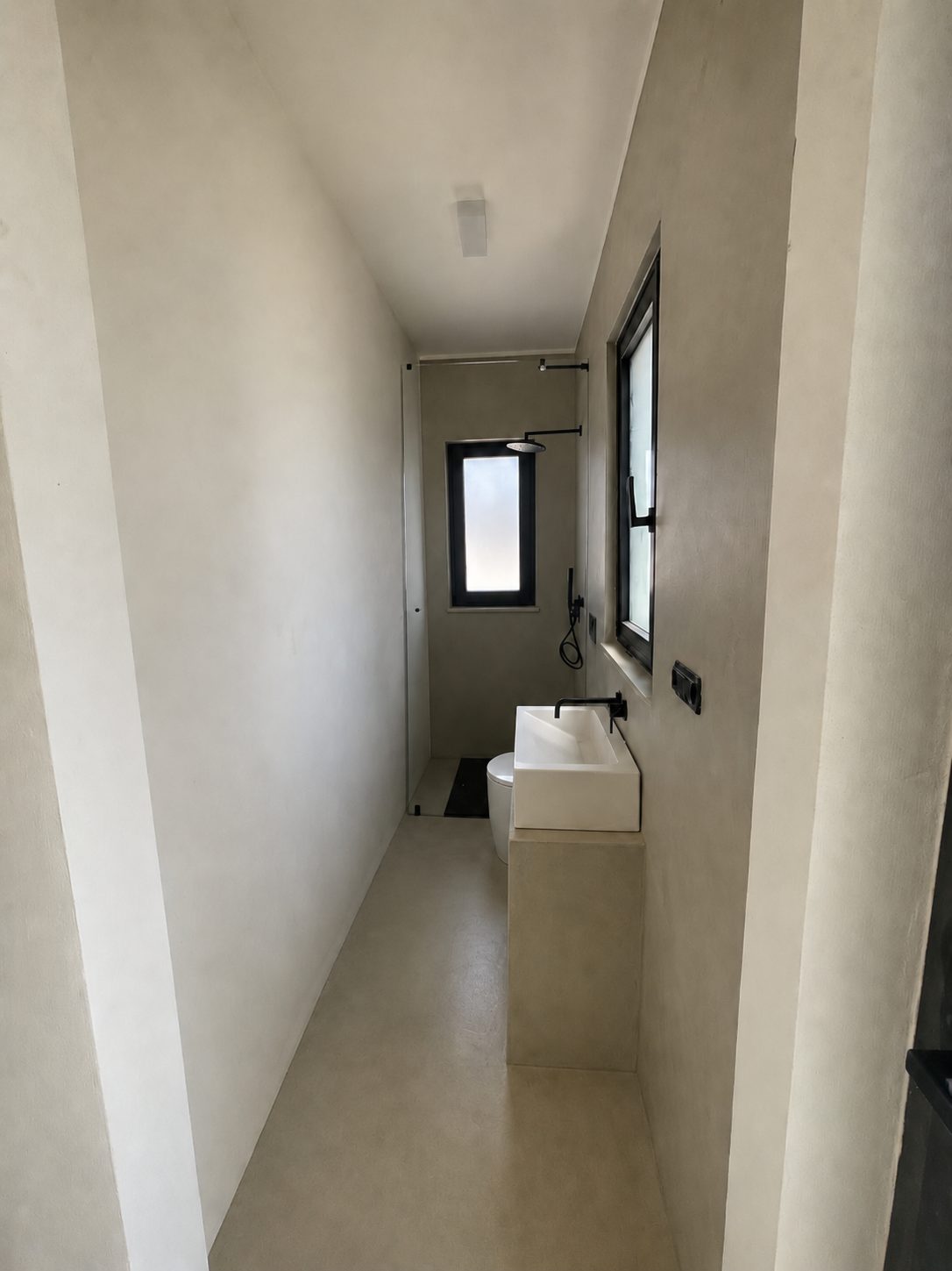 La salle de bains des invités de Casa Andrea — pièce étroite en microciment d'un ton chaud, vasque maçonnée et robinetterie murale noire, fenêtre à cadre noir, douche à l'italienne au fond, Pouilles.