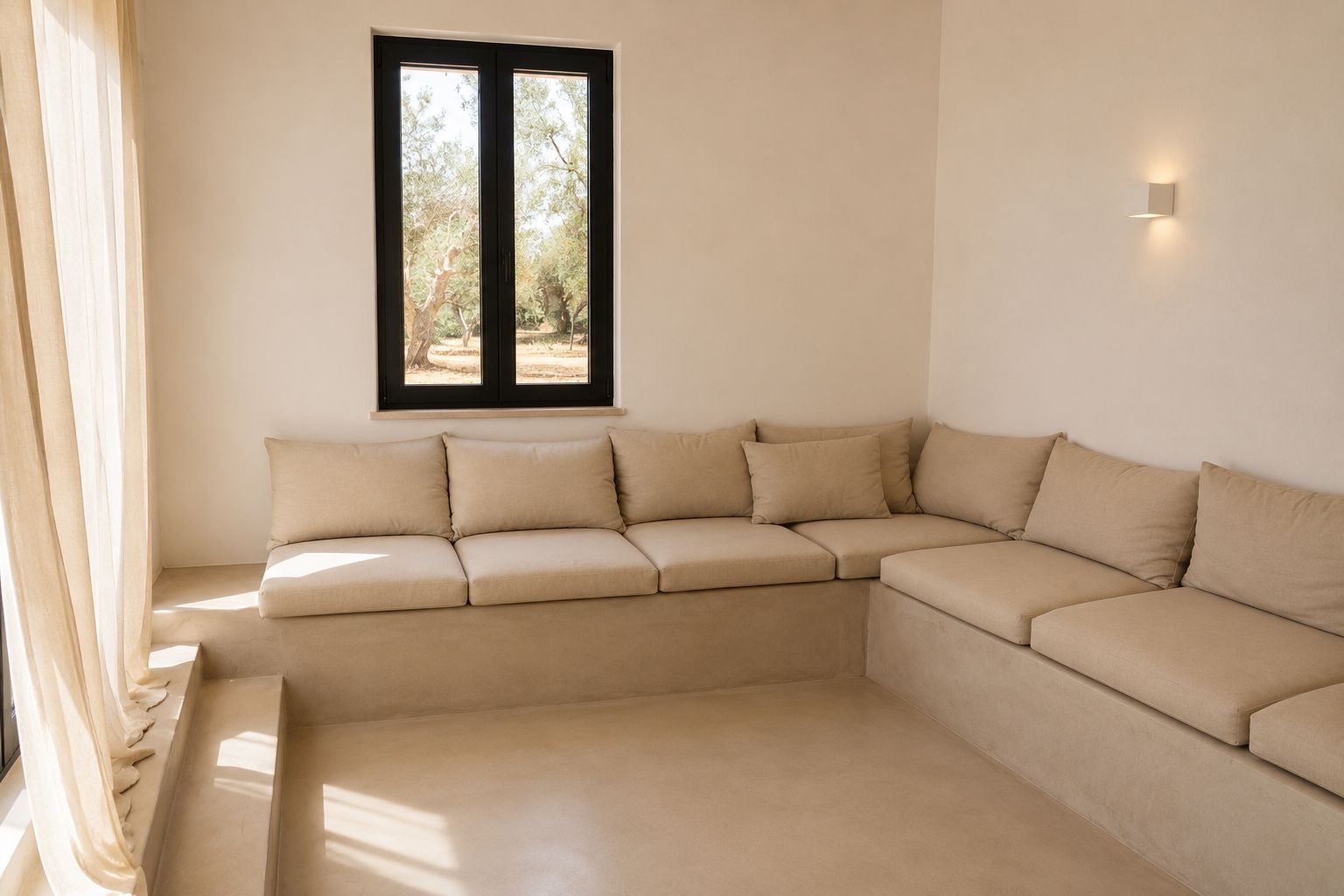 La banquette du salon de Casa Andrea — une longue assise en microciment le long de deux murs, coussins de lin, une fenêtre à cadre noir donnant droit sur les oliviers, une applique en plâtre, le soleil bas sur le sol, Pouilles.