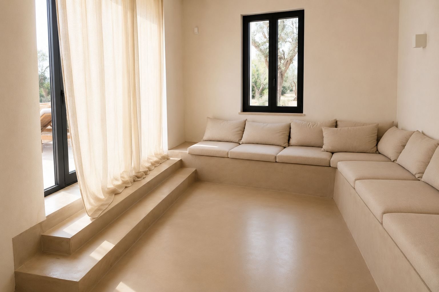 Le salon de Casa Andrea — une longue banquette maçonnée en microciment garnie de coussins de lin le long de deux murs, une fenêtre à cadre noir sur un olivier, un rideau de lin à la porte vitrée, deux marches basses dans le sol, Pouilles.