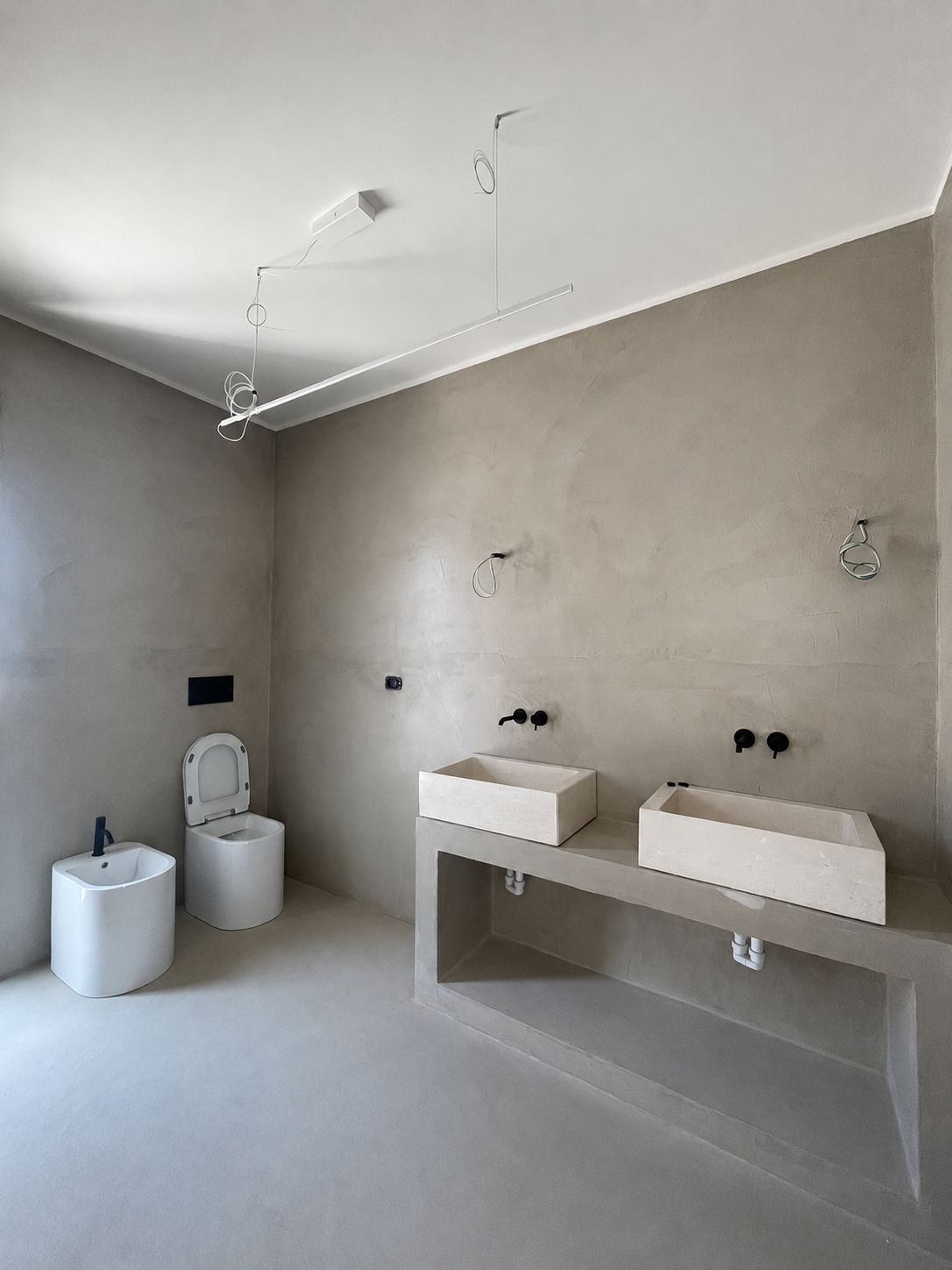 La salle de bains attenante de la master de Casa Andrea — un double meuble-lavabo en microciment à deux vasques et robinetterie murale noire, un bidet et un WC, murs et sol d'un même ton chaud, Pouilles.