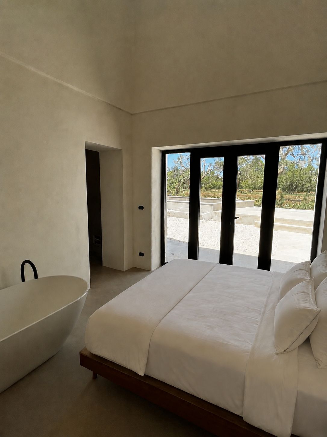 La master bedroom sous un autre angle — une baignoire sculpturale près du lit, de grandes baies vitrées toute hauteur à cadre noir ouvertes sur l'oliveraie, sol en microciment, Pouilles.