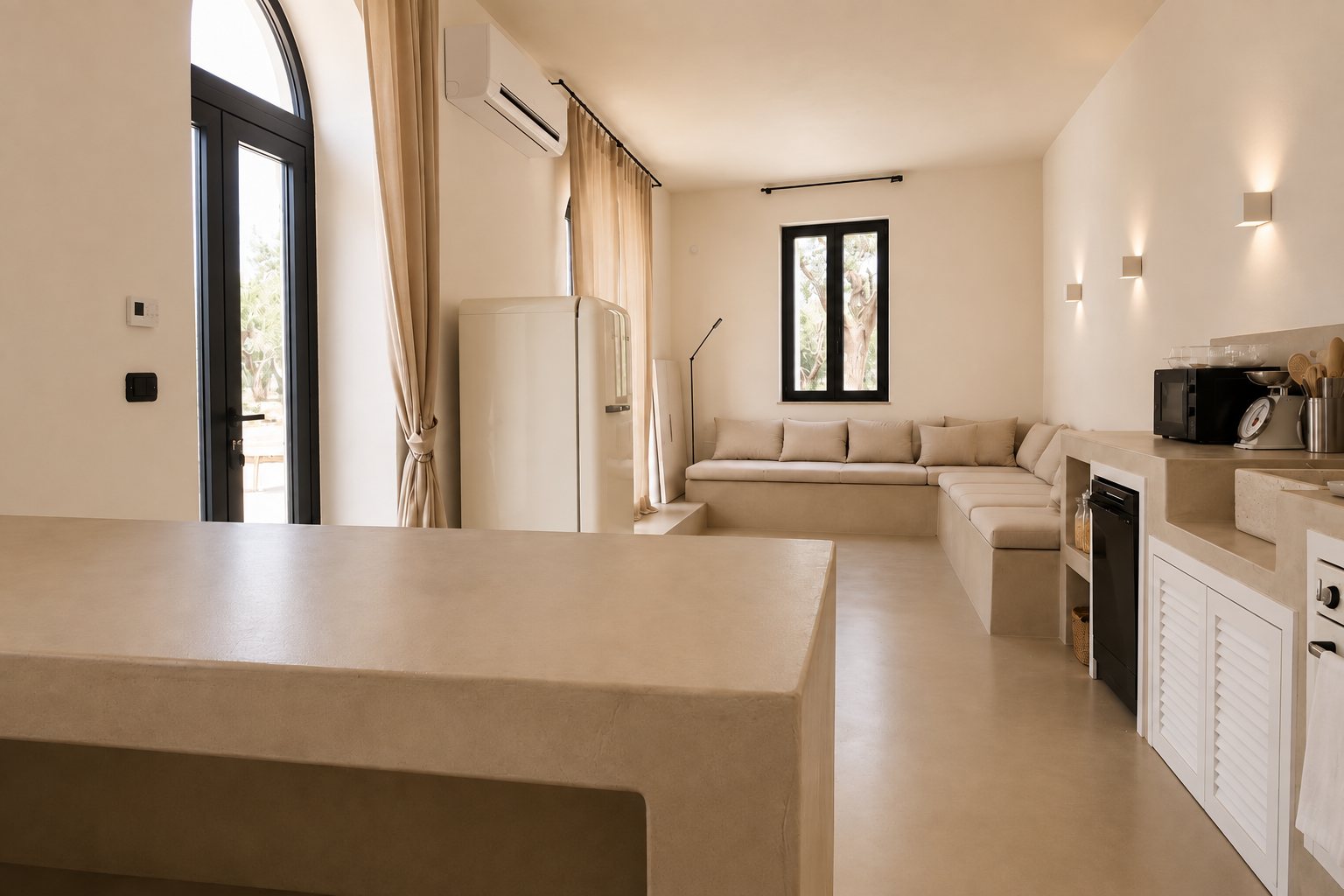 Le salon ouvert et la cuisine de Casa Andrea vus depuis l'îlot — la haute porte cintrée vitrée et son rideau de lin d'un côté, un réfrigérateur crème, la banquette maçonnée sous une fenêtre sur les oliviers, le plan de cuisine et ses appliques de plâtre de l'autre, microciment partout, Pouilles.