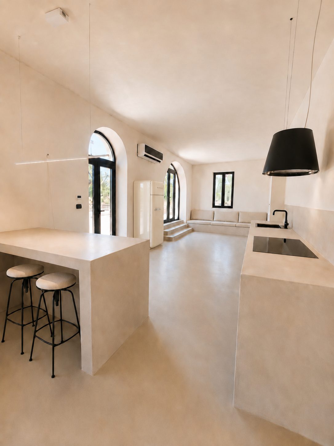 Le salon ouvert et la cuisine de Casa Andrea — un bar en pierre avec étagère ouverte pour les tabourets à venir, le plan de cuisine le long du mur, deux fenêtres cintrées vitrées sur la cour, une suspension noire, sol en microciment, Pouilles.