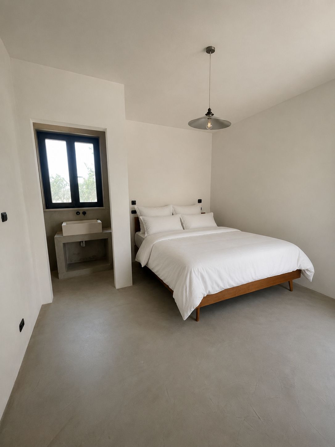 La troisième chambre en suite de Casa Andrea — murs et sol en microciment d'un ton chaud, lit en bois et linge blanc, une ouverture vers la salle d'eau attenante et sa vasque, Pouilles.