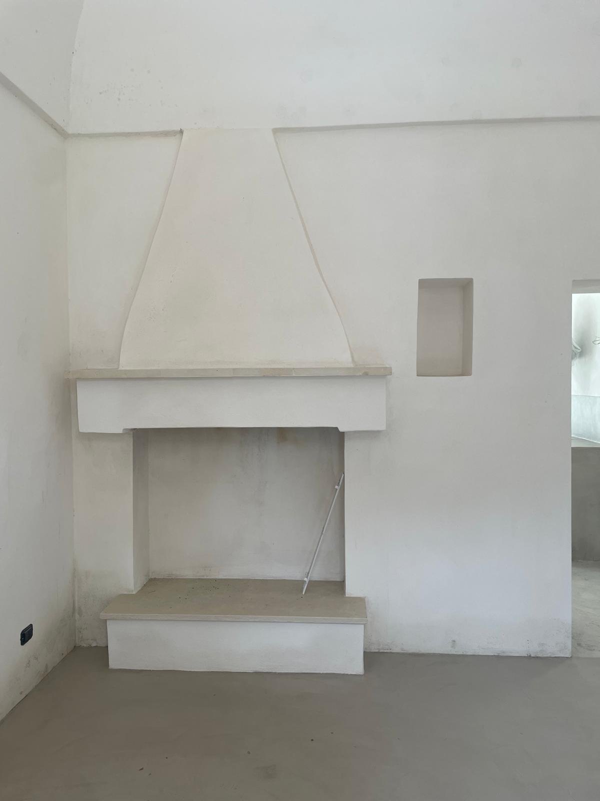 La cheminée restaurée du casale de Casa Andrea, mars 2026 — cappa pugliese conique en plâtre blanc, linteau et foyer en Pietra di Trani, banquette panca camino intégrée, niche sculptée dans le mur.